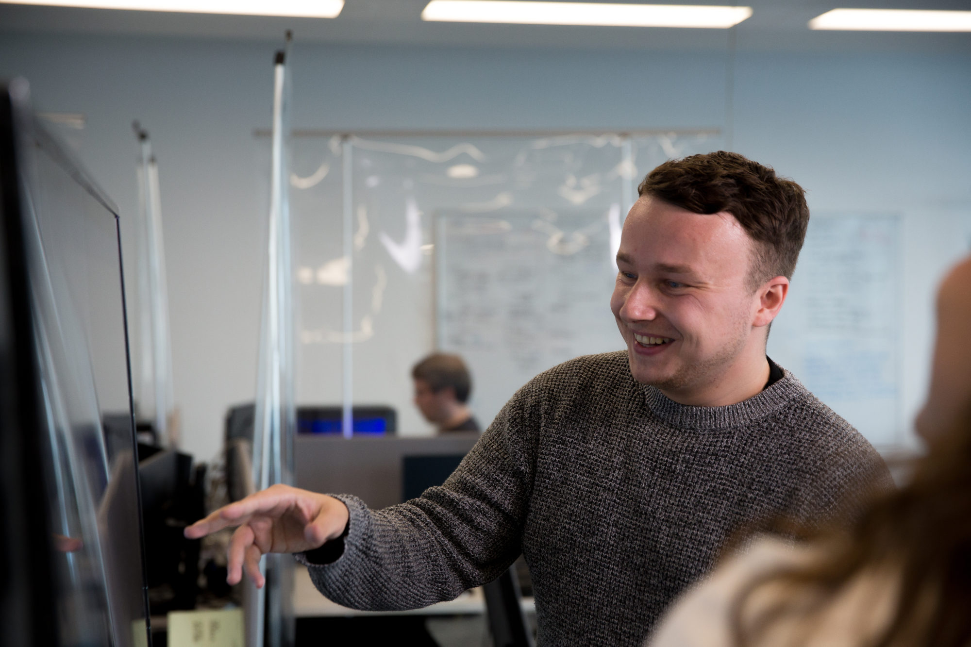 A male coder of a digital twin company smiling while pointing to a television screen and explaining to another female employee.