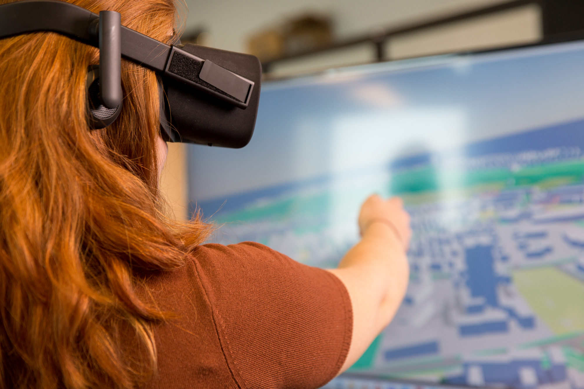A female employee of Slingshot Simulations wearing a VR headset and pointing at a screen displaying 3D renders.