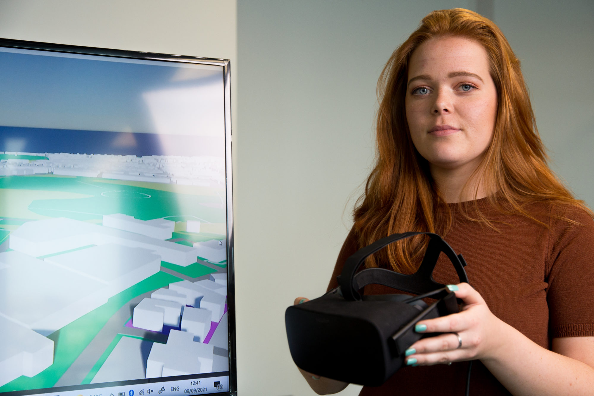 A female Slingshot Simulations employee holding a VR headset and standing in front of a TV screen displaying 3D renders.