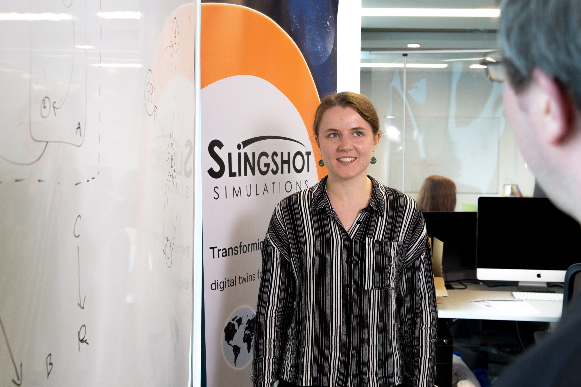 A female member of the Slingshot Simulations team standing in front of a company banner smiling and chatting to a male employee.